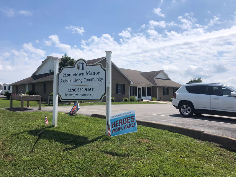 Exterior view of Hometown Manor Assisted Living Community building with a large white sign displaying the facility name, phone number, and website. A white SUV is parked in the parking lot beside the building. There is a smaller sign in the grass that reads 'Heroes Work Here!' with small American flags nearby. The sky is partly cloudy.