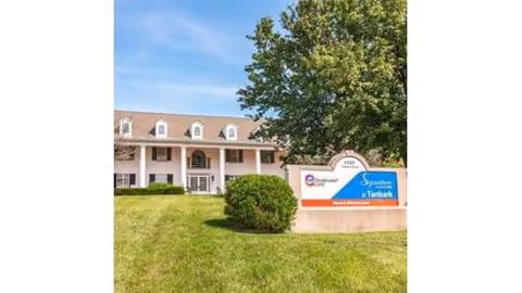 Exterior view of Signature HealthCARE at Tanbark Rehab & Wellness Center showing a two-story building with white columns and dormer windows, a large tree, green lawn, and a sign with the facility's name in front.
