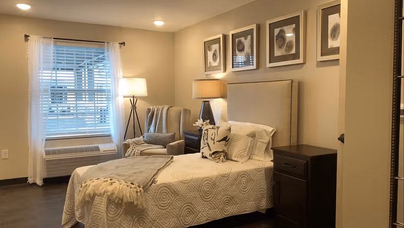 Furnished bedroom with a single bed, upholstered headboard, bedside tables and lamps, an armchair by a window with white blinds, and neutral decor.