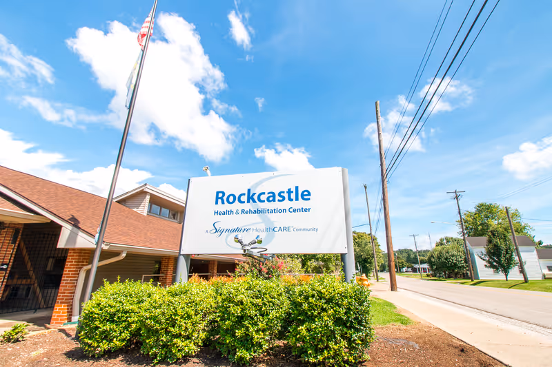 Exterior view of the Rockcastle Health & Rehabilitation Center sign and entrance along a street under a blue sky.