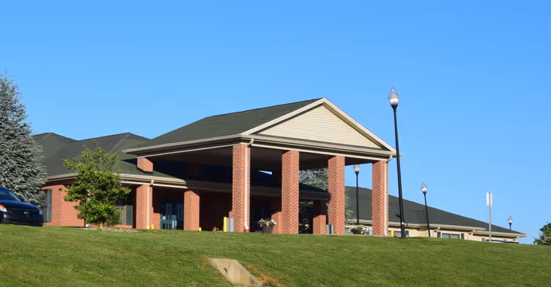 Front entrance of a single-story brick senior living facility with a covered porte-cochère, lamp posts, and a grassy lawn under a clear blue sky.