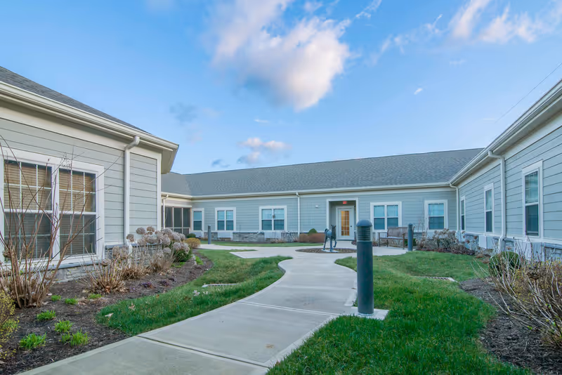 Courtyard walkway between single-story Morning Pointe of Lexington buildings with landscaping, benches and a small deer sculpture under a blue sky.