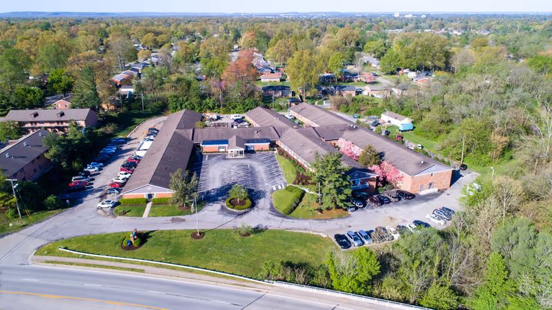 Aerial view of Hillcreek Rehabilitation and Care facility showing multiple connected buildings with brown roofs surrounded by trees and greenery. There are parking lots with several cars parked and a main road in the foreground.
