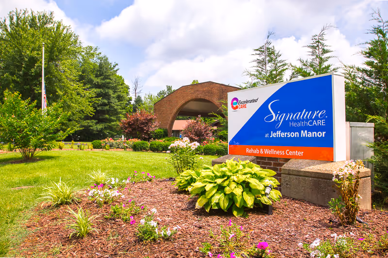 Outdoor view of the entrance area of Signature HealthCARE at Jefferson Manor Rehab & Wellness Center, featuring a landscaped garden with green plants and flowers, a large sign with the facility's name, and a brick archway in the background under a partly cloudy sky.