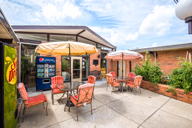 Outdoor patio area with two glass-top tables, each surrounded by four cushioned chairs with red and orange patterned upholstery. Each table has a large umbrella for shade. There are vending machines for Dr Pepper and Pepsi against a brick wall near glass doors leading inside. Raised brick planters with green plants are visible on the right side. The sky is partly cloudy.