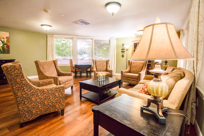 A cozy living room with a beige sofa, four patterned armchairs arranged around a dark wooden coffee table, a side table with a large lamp, wooden flooring, and large windows with curtains letting in natural light.