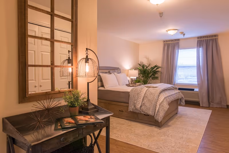 Cozy furnished bedroom with a bed, bedside tables and lamps, a decorative mirror, desk with decor, and a window with curtains.