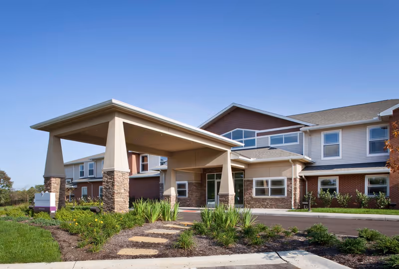 Exterior view of a modern senior living facility building with a covered entrance supported by stone and beige pillars, surrounded by landscaped greenery and a clear blue sky.