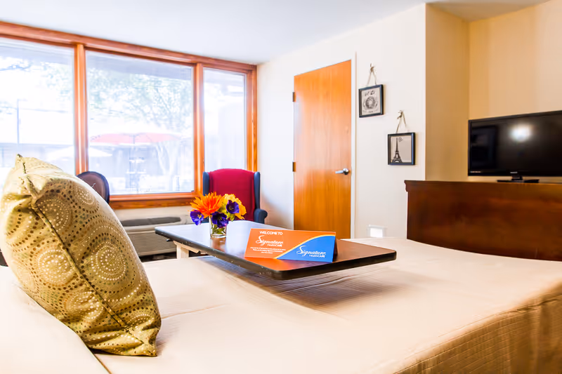 A bright and cozy room with a large window letting in natural light. The room features a bed with a decorative pillow, a small table with a vase of colorful flowers and a welcome card from Signature HealthCARE. There is a wooden door, two framed pictures on the wall, a red armchair, and a flat-screen TV on a wooden stand.