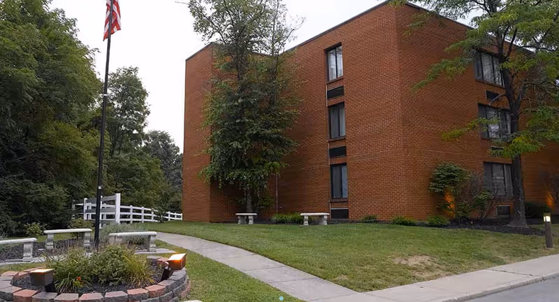 Exterior view of a brick building with several windows, surrounded by green trees and grass. A paved walkway curves through the lawn, leading past stone benches and a flagpole with an American flag. The scene appears to be part of a senior living facility.