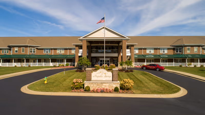 Front exterior view of Magnolia Springs Lexington, a large two-story senior living facility with a central entrance, an American flag on a flagpole, manicured lawns, and a driveway with a red car parked on the right side.