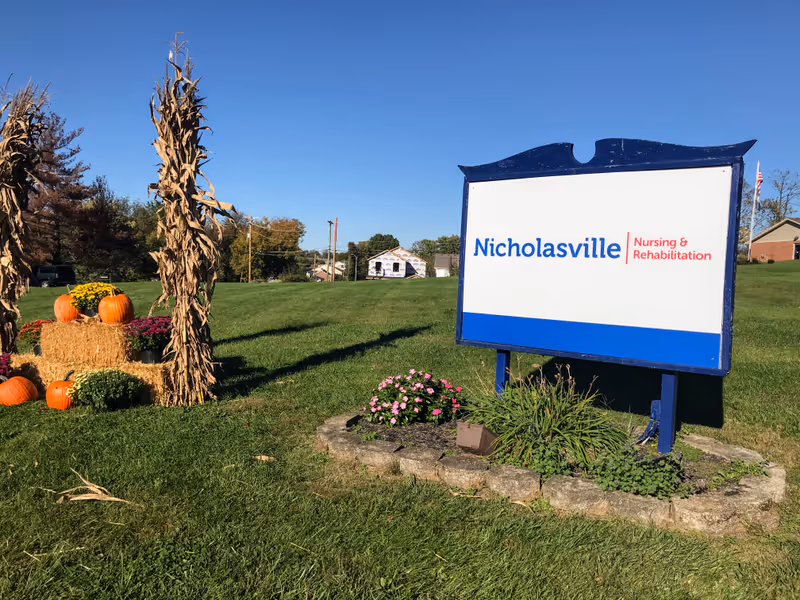 Outdoor scene showing a large sign for Nicholasville Nursing & Rehabilitation on a grassy lawn with a clear blue sky. To the left of the sign, there is a fall-themed decoration with hay bales, pumpkins, flowers, and dried cornstalks. Trees and houses are visible in the background.
