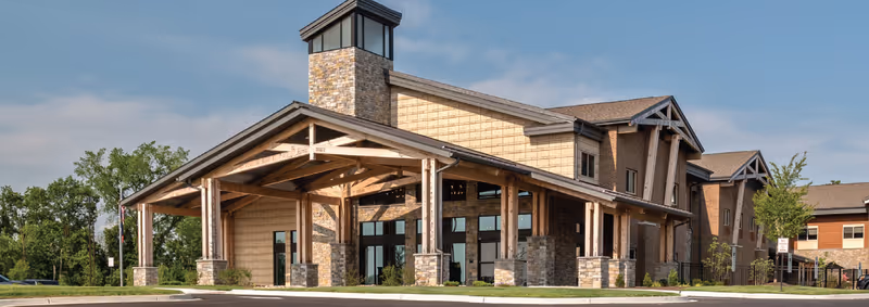 Exterior view of Boonespring Skilled Nursing Facility showing a large building with a covered entrance supported by wooden beams and stone pillars, surrounded by greenery and a clear sky.