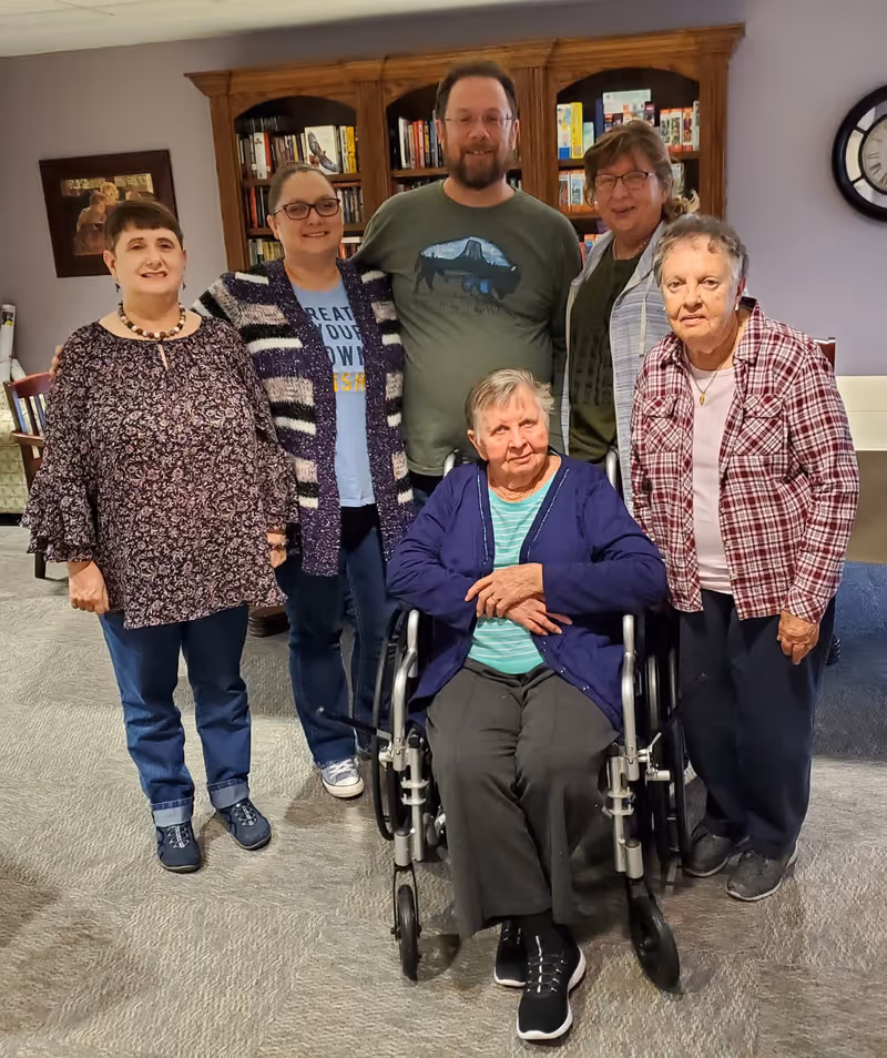 Group of six adults, including one in a wheelchair, posing in a library-style common area.