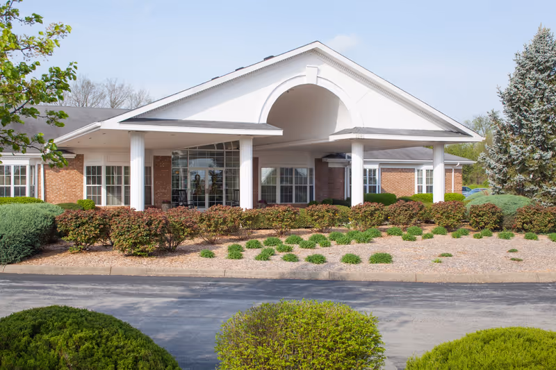 Front exterior view of a single-story brick building with white columns and a covered entrance, surrounded by landscaped bushes and trees under a clear sky.