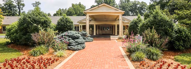 Front exterior view of Friendship Health & Rehab building with a covered entrance, brick walkway, and landscaped garden beds with various shrubs and flowers on either side.