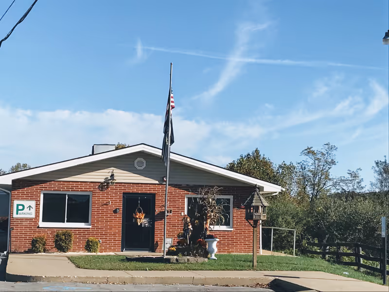 Front exterior view of a single-story brick building with a triangular roof, two windows, a door decorated with autumn wreaths, a flagpole with flags, and a small garden area with bushes and a birdhouse. The sky is clear and blue.