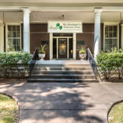 Front entrance of The Altenheim Senior Health Care Community featuring a short staircase flanked by railings and columns, potted plants, and a sign above the doorway.