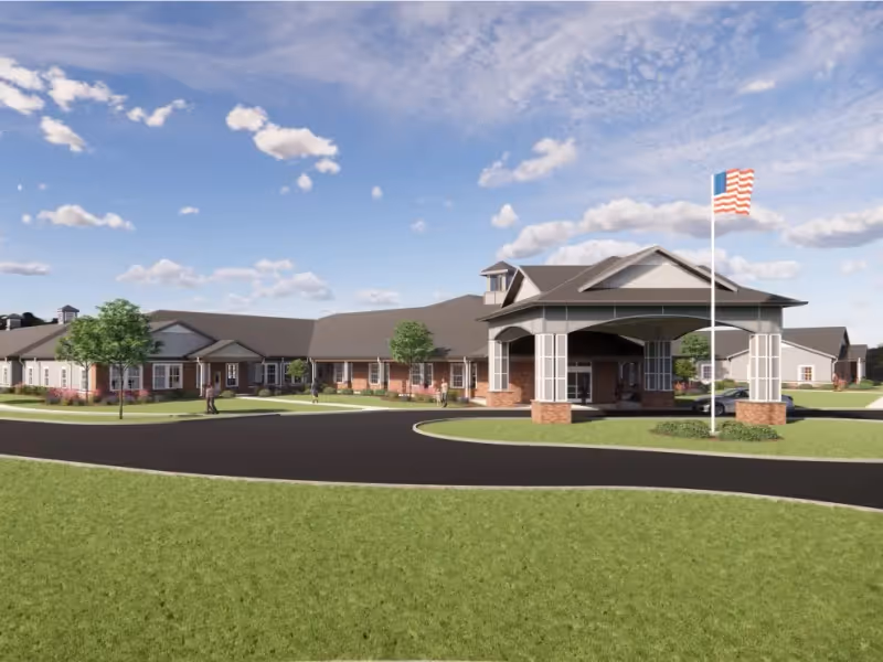 Exterior front view of a single-story assisted living building with a covered porte-cochere, American flag, and landscaped lawn.