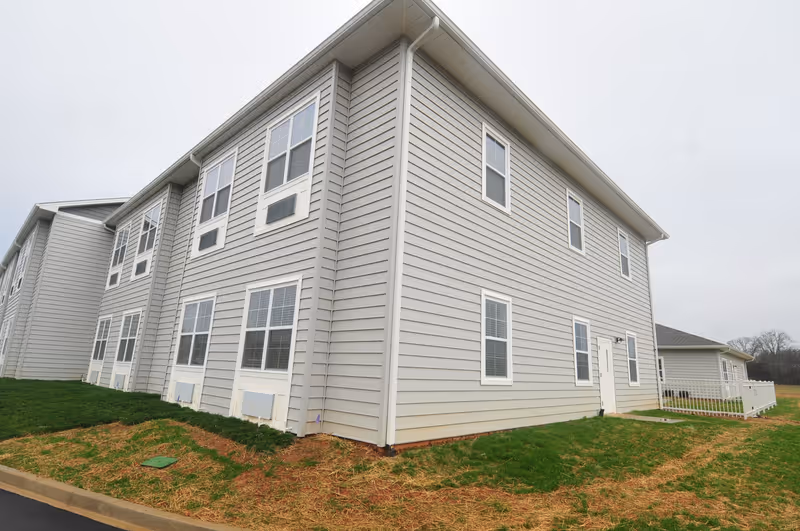 Exterior view of a two-story gray building with multiple windows and a white door, surrounded by grass and a paved area under an overcast sky.