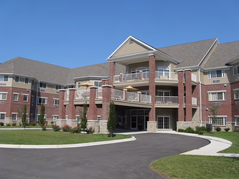 Exterior view of a large senior living facility building with brick and beige siding, multiple windows, balconies with railings and umbrellas, a driveway, and landscaped greenery under a clear blue sky.