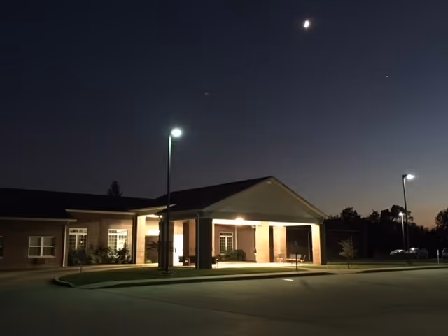 Exterior view of Breckinridge Place Retirement Community at night, showing a single-story building with a covered entrance illuminated by outdoor lights under a dark sky with a crescent moon visible.