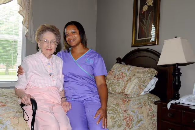 An elderly woman sitting on the edge of a bed holding a cane, accompanied by a smiling caregiver in purple scrubs sitting beside her in a bedroom with floral bedding, a bedside table with a lamp and a phone, and a window with blinds and curtains.