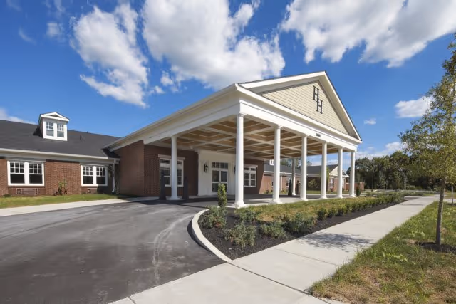 Exterior view of Hallmark House, a single-story brick building with a large covered entrance supported by white columns. The sky is blue with scattered clouds, and there is a sidewalk and landscaped greenery in front of the building.