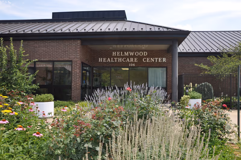 Entrance of Helmwood Healthcare Center with a brick exterior, large windows, and a metal roof. In front of the building is a garden with various flowering plants and greenery.
