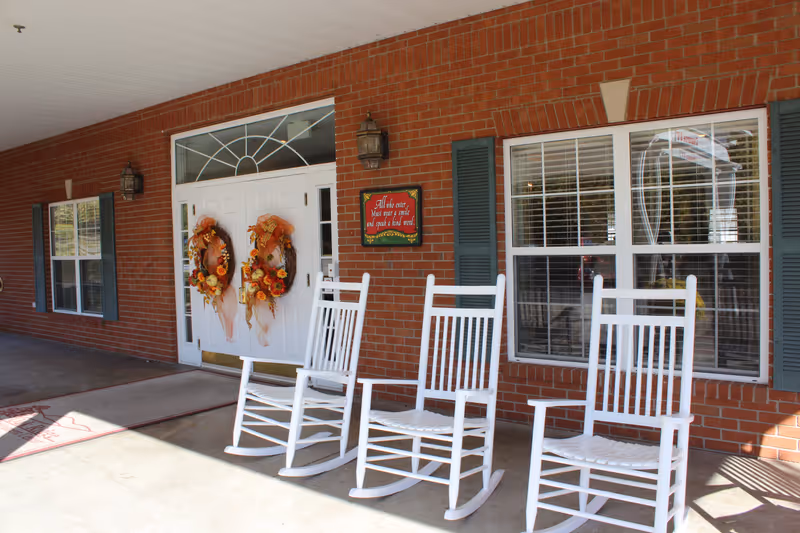 Front porch area of a senior living community with three white rocking chairs, a brick wall, two windows with green shutters, and double white doors decorated with autumn-themed wreaths. A small sign with a welcoming message is mounted on the wall between the doors and the window.