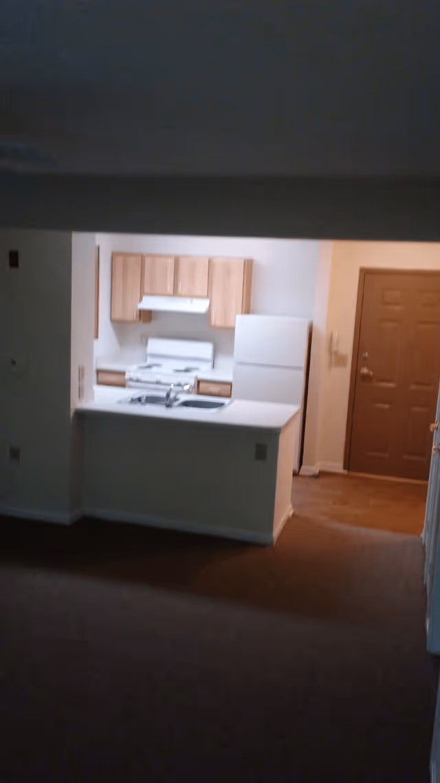 View of a small kitchen area in an apartment with wooden cabinets, a white stove with a range hood, a white refrigerator, and a countertop with a sink. The kitchen opens to a carpeted living space and there is a wooden door visible to the right.