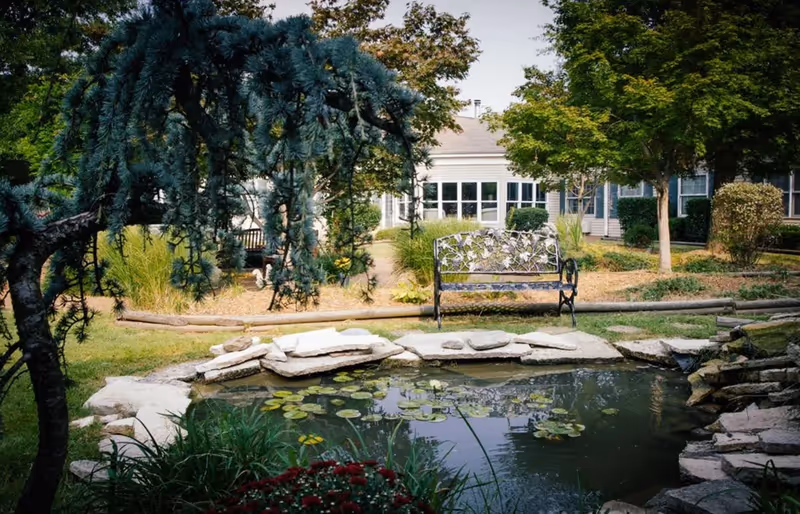 Tranquil outdoor garden at a senior living facility with a pond, bench, and stone edging.