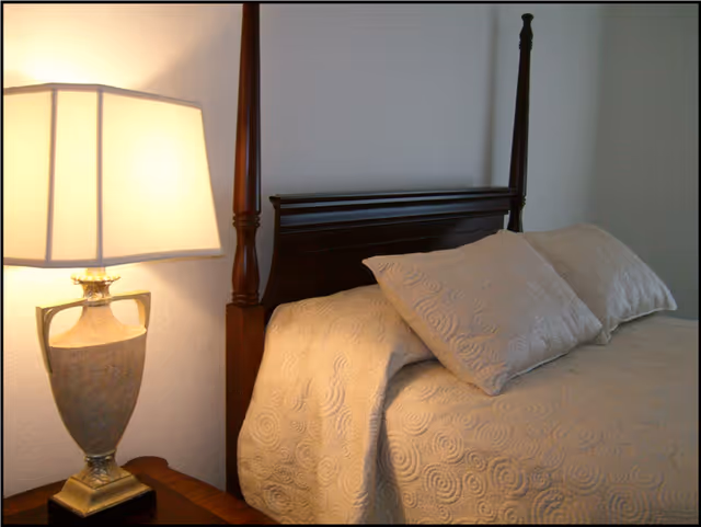 Well-lit bedroom with a wooden four-poster headboard, beige patterned bedspread and pillows, and a table lamp on a nightstand.