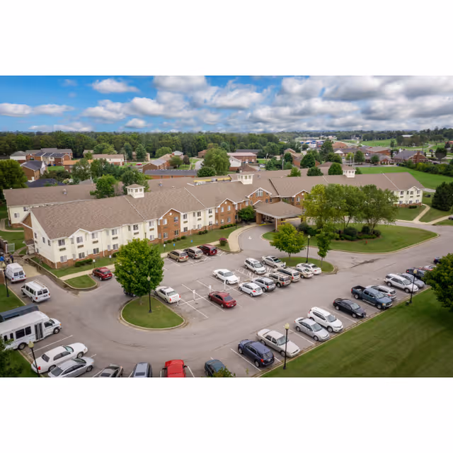 Aerial view of McDowell Place of Danville, showing a large two-story building with a beige and brick exterior surrounded by a parking lot with numerous cars. The building has a covered entrance and is set in a green landscape with trees and a partly cloudy sky overhead.
