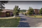 View of a paved road leading into a senior living community with brick buildings on either side, green lawns, and several trees under a clear sky.