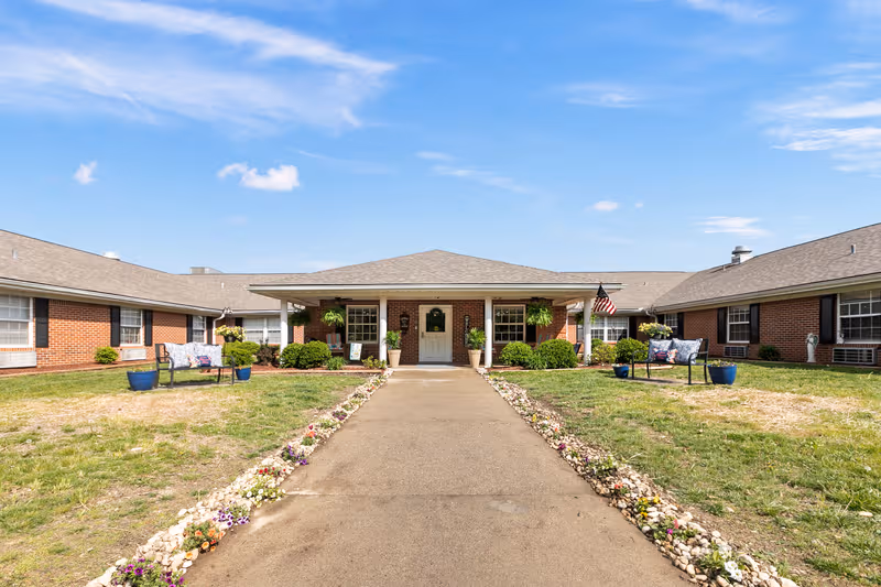Oakmont Manor exterior: brick building with central entrance, flower-lined walkway, and benches.
