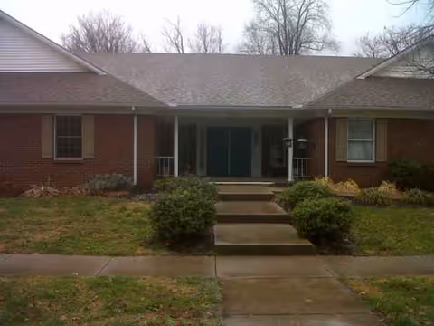 Brick single-story assisted living facility with central entrance, steps, and lawn-lined path.
