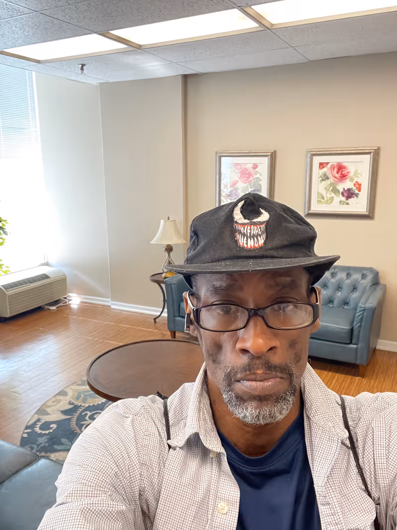 Selfie of a man wearing a hat in a lounge with blue couch and coffee table.