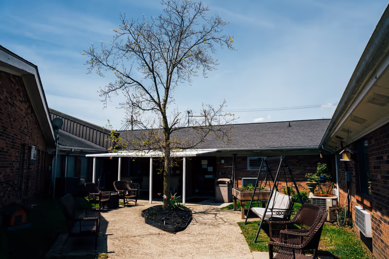 Outdoor courtyard area at Frankfort Trails featuring a central tree with budding leaves, surrounded by a paved walkway and multiple seating options including chairs and a swing. The courtyard is enclosed by brick buildings under a clear blue sky.