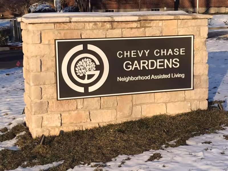 Stone sign reads Chevy Chase Gardens Neighborhood Assisted Living in an outdoor setting.