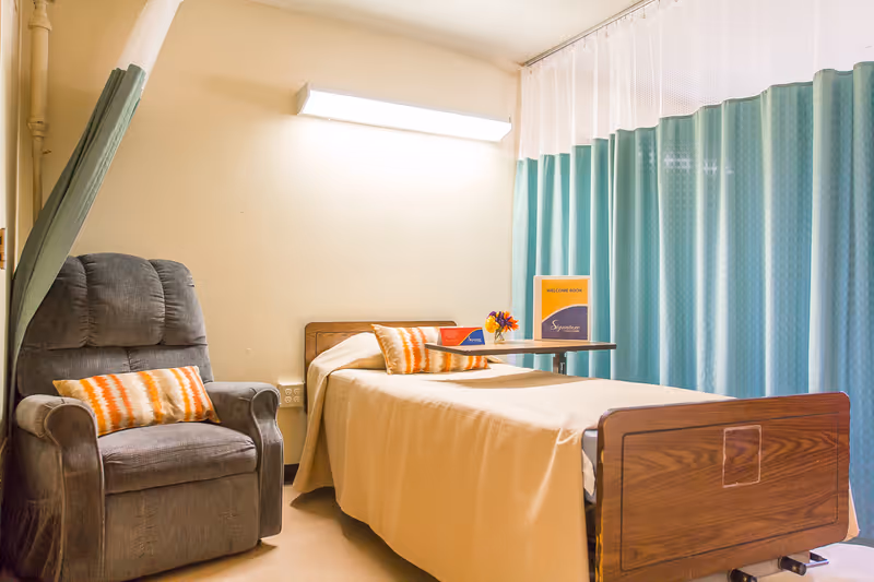 A small rehab room with a single hospital bed, a gray recliner with patterned pillows, an overbed table, and teal privacy curtains.