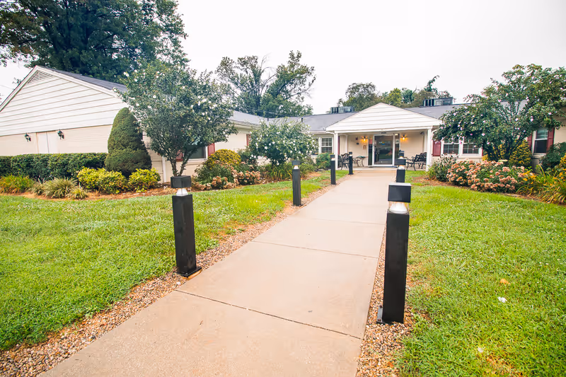 Front entrance of a single-story senior living facility with a walkway lined by low light posts and landscaped lawns.