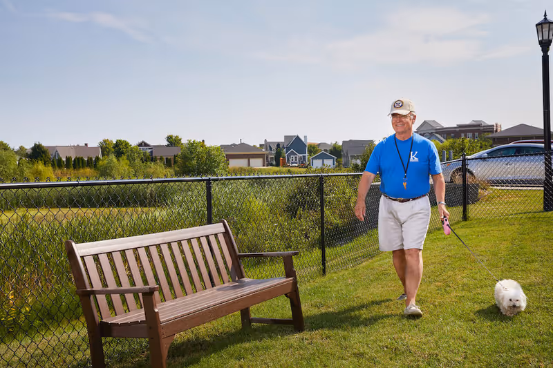 An elderly man wearing a blue shirt, beige shorts, and a beige cap is walking a small white dog on a leash along a grassy area next to a chain-link fence. There is a wooden bench nearby, and houses and trees are visible in the background under a partly cloudy sky.
