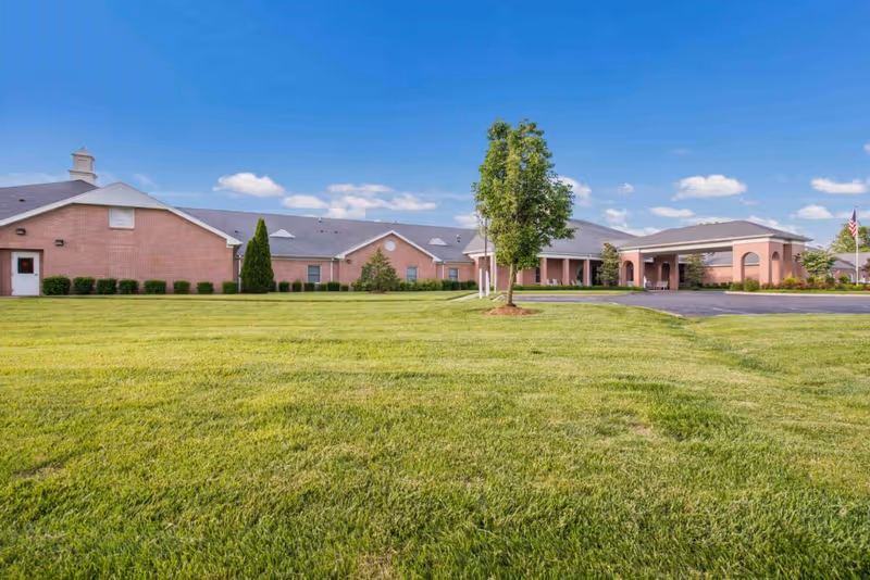 Exterior view of a single-story brick building with a well-maintained lawn, a few trees, and a clear blue sky with some clouds. The building has a covered entrance with columns and an American flag is visible on the right side near the driveway.