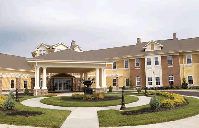 Exterior view of a senior living community building with a covered entrance supported by white columns. The building has a combination of yellow siding and brick facade with multiple windows. There is a circular driveway with landscaped greenery and a decorative urn in the center.