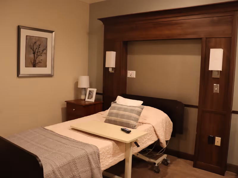 A single bed with a beige quilt and a gray plaid pillow in a room with beige walls. The bed has a small overbed table with a remote control on it. There is a wooden nightstand with a lamp and a framed photo, and a framed artwork of a tree branch on the wall. The bed is set against a wooden headboard with built-in wall lights.