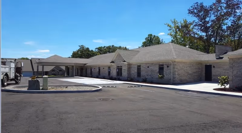 Exterior view of a single-story brick building with a covered entrance and a paved driveway under a clear blue sky. Trees are visible in the background.