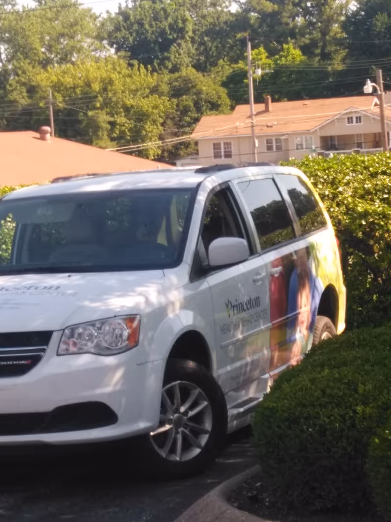 White Princeton Health & Rehab van parked beside greenery with a residential building behind.