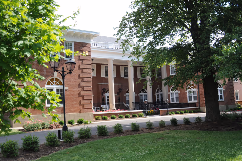 Exterior view of a senior living facility building with red brick walls, white columns, and multiple windows. The foreground features a well-maintained lawn, small bushes, a large tree, and a black street lamp.