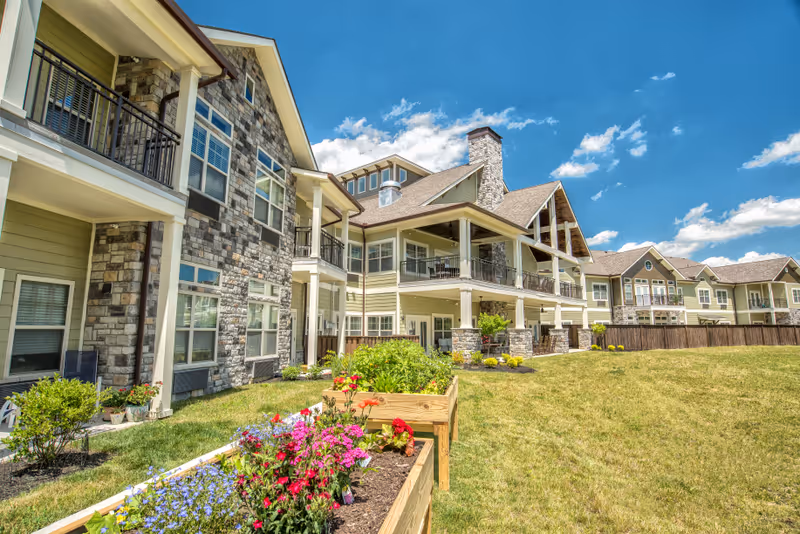 Exterior view of a multi-story senior living facility with stone accents, covered balconies, and a grassy lawn with raised flower beds under a blue sky.